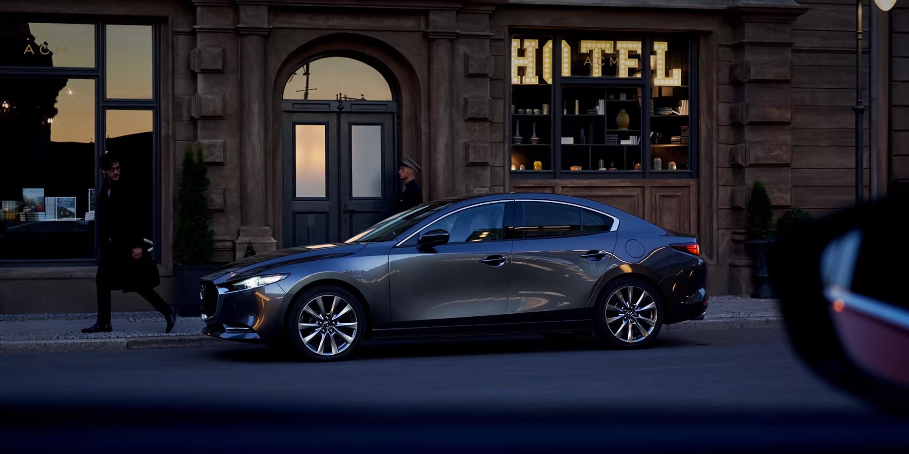 Gray sedan parked on a city street at dusk with pedestrians walking by.