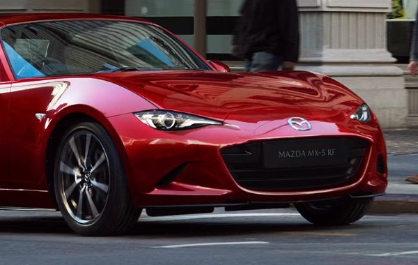 Red Mazda MX-5 car in motion on city street with pedestrians.